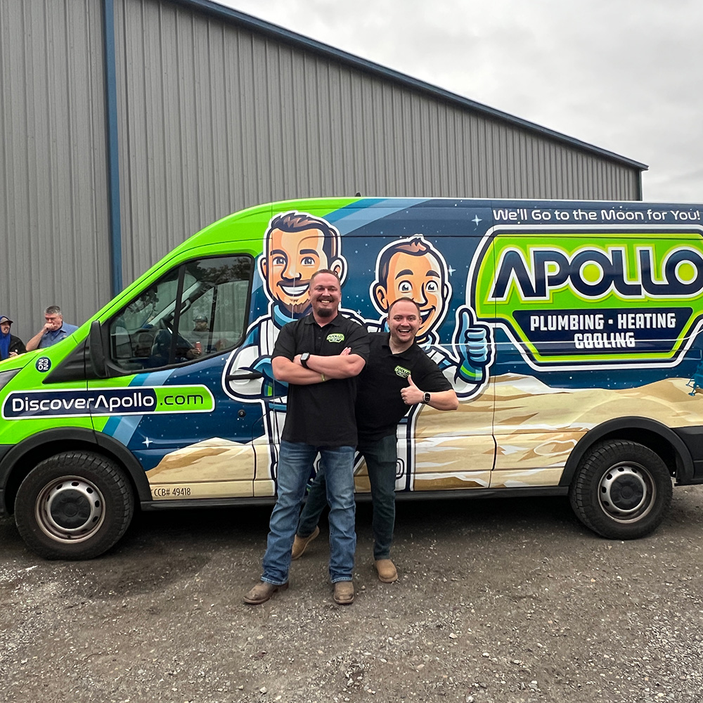 Two smiling men wearing black Apollo shirts link arms and stand before a bright Apollo Plumbing, Heating, Cooling van with cartoons.