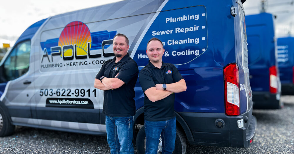 Two smiling individuals in polo shirts stand back-to-back in front of a blue plumbing service van that displays contact information and a list of services.